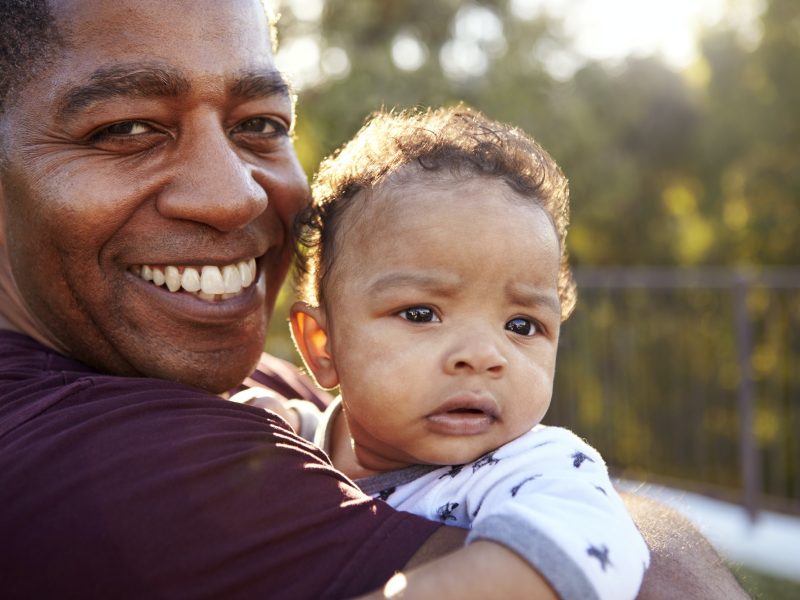 Grandfather standing in the garden holding his three month old grandson, smiling